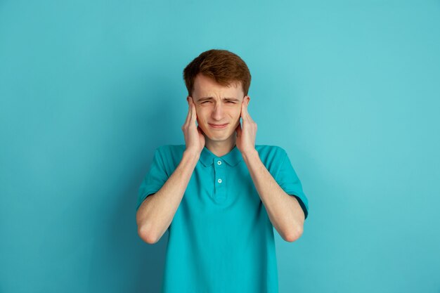 Dolor de cabeza, malestar. Retrato moderno del hombre joven caucásico aislado en la pared azul, monocromo. Hermoso modelo masculino.