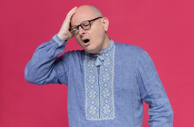 Dolor de hombre eslavo adulto en camisa azul con gafas ópticas poniendo la mano sobre su cabeza