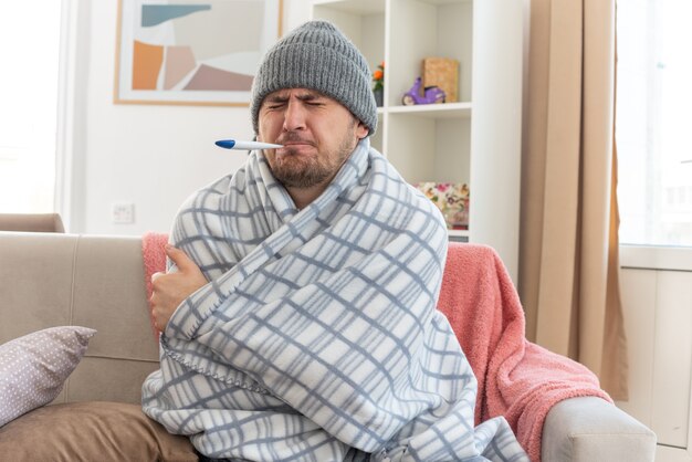 Dolor de hombre eslavo enfermo con bufanda alrededor del cuello con sombrero de invierno envuelto en cuadros midiendo su temperatura con termómetro sentado en el sofá en la sala de estar