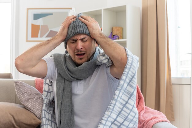 Dolor de hombre eslavo enfermo con bufanda alrededor del cuello con sombrero de invierno envuelto en cuadros sosteniendo su cabeza sentado en el sofá en la sala de estar