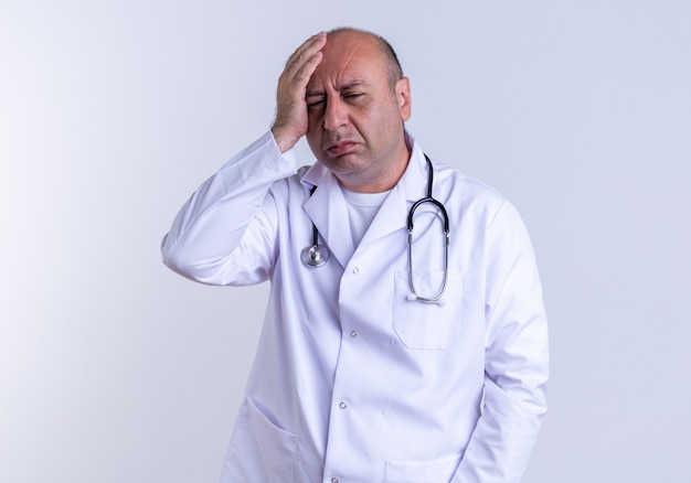 Dolor de hombre de mediana edad médico vestido con bata médica y estetoscopio manteniendo la mano en la cabeza mirando hacia abajo aislado en la pared blanca