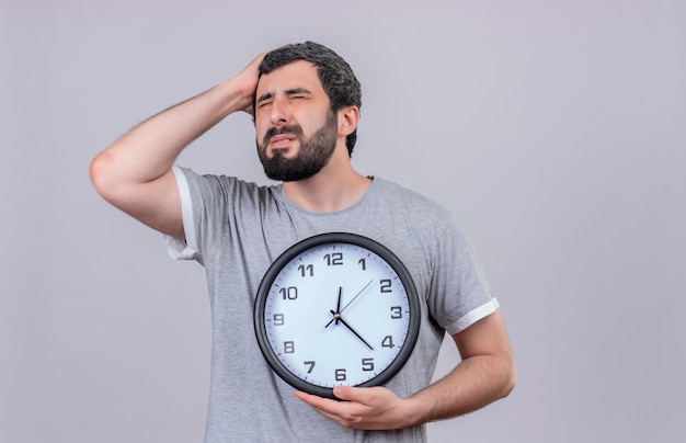 Dolor joven apuesto hombre caucásico que sostiene el reloj poniendo la mano en la cabeza que sufre de dolor con los ojos cerrados aislado en blanco con espacio de copia