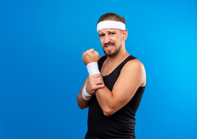 Dolor joven apuesto hombre deportivo con diadema y muñequeras apretando el puño y sosteniendo su brazo aislado en la pared azul con espacio de copia