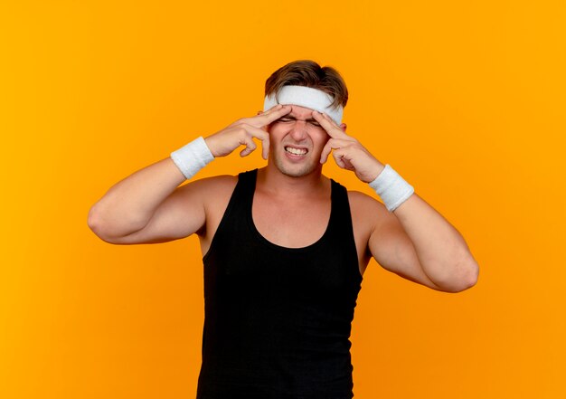 Dolor joven apuesto hombre deportivo con diadema y muñequeras poniendo los dedos en la frente que sufre de dolor de cabeza aislado en naranja