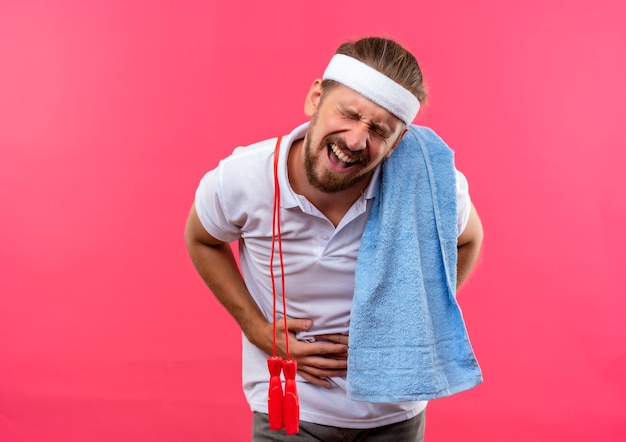 Foto gratuita dolor joven apuesto hombre deportivo con diadema y muñequeras sosteniendo su vientre con los ojos cerrados con saltar la cuerda y una toalla sobre sus hombros aislado en la pared rosa con espacio de copia