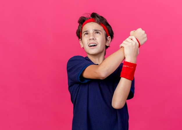 Foto gratuita dolor joven apuesto muchacho deportivo con diadema y muñequeras con aparatos dentales mirando hacia arriba sosteniendo la muñeca aislada en la pared carmesí