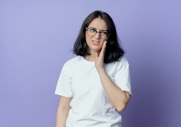 Dolor joven bastante caucásica con gafas poniendo la mano en la mejilla mirando al lado que sufre de dolor de muelas aislado sobre fondo púrpura con espacio de copia