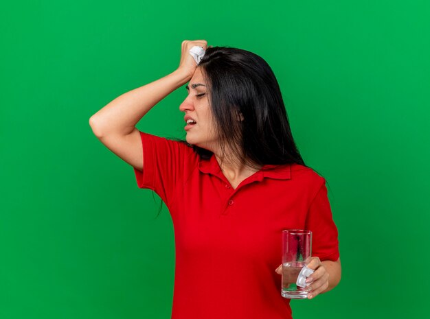 Dolor a joven caucásica enferma sosteniendo paquete de tabletas vaso de agua y servilleta girando la cabeza a un lado manteniendo la mano en la cabeza que sufre de dolor de cabeza aislado sobre fondo verde con espacio de copia