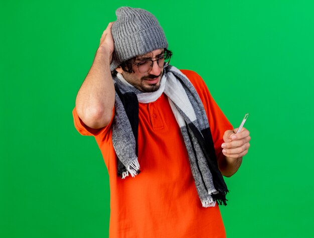Dolor joven caucásico enfermo con gafas sombrero de invierno y bufanda sosteniendo el termómetro manteniendo la mano en la cabeza aislada sobre fondo verde con espacio de copia