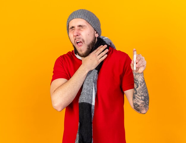 Dolor joven caucásico enfermo vistiendo gorro y bufanda de invierno pone la mano en el cuello y sostiene el termómetro