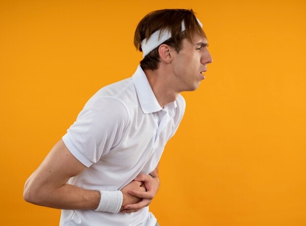 Dolor joven deportista con diadema con muñequera agarró el estómago aislado en la pared amarilla