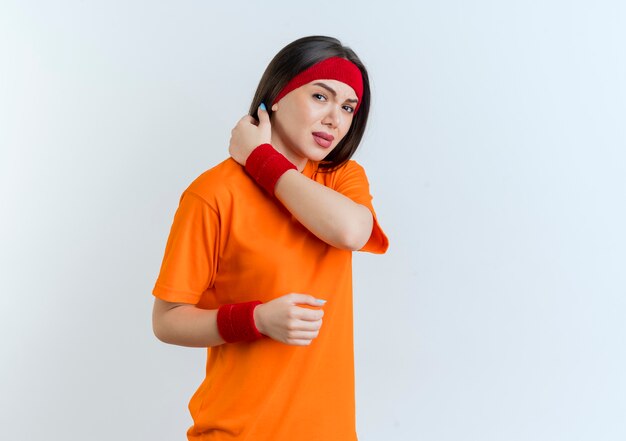 Dolor de joven deportiva vistiendo diadema y muñequeras mirando manteniendo la mano en el aire tocando el cuello aislado