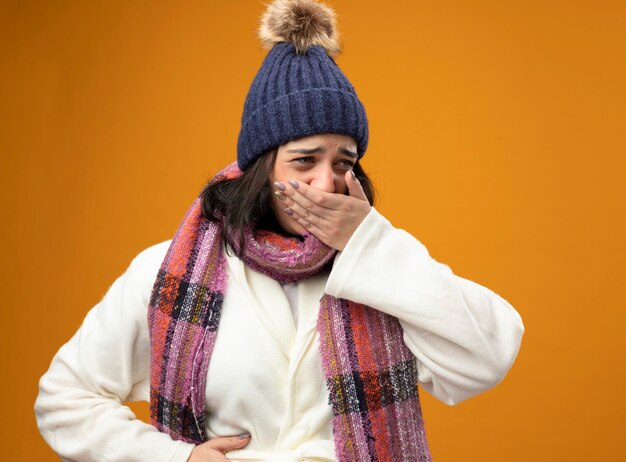 Dolor a joven enferma caucásica vistiendo bata sombrero de invierno y bufanda con náuseas poniendo la mano en el vientre y en la boca mirando al lado aislado en la pared naranja