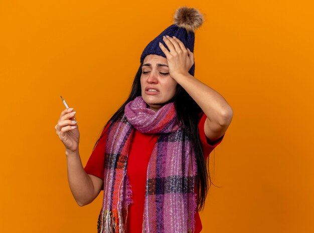 Dolor de joven enferma caucásica vistiendo gorro y bufanda de invierno sosteniendo y mirando el termómetro aislado en la pared naranja con espacio de copia