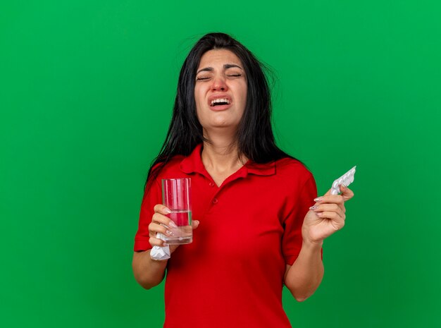 Dolor de joven enferma sosteniendo un paquete de tabletas vaso de agua y una servilleta con los ojos cerrados aislado en la pared verde