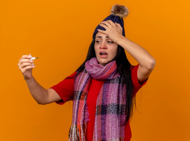 Foto gratuita dolor de joven enferma vistiendo gorro de invierno y bufanda sosteniendo y mirando el termómetro poniendo la mano en la cabeza aislada en la pared naranja
