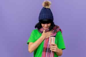 Foto gratuita dolor de joven enferma vistiendo gorro de invierno y bufanda sosteniendo un paquete de cápsulas manteniendo la mano en el pecho con los ojos cerrados aislados en la pared púrpura