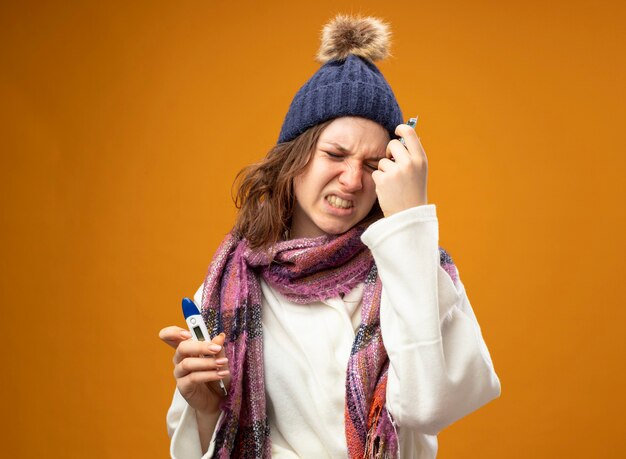 Dolor a joven enferma vistiendo túnica blanca y gorro de invierno con bufanda sosteniendo el termómetro con pastillas poniendo la mano en la frente aislada en la pared naranja