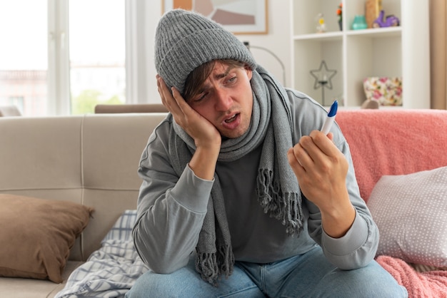 Foto gratuita dolor joven enfermo con bufanda alrededor del cuello con sombrero de invierno poniendo la mano en la cara sosteniendo y mirando el termómetro sentado en el sofá en la sala de estar