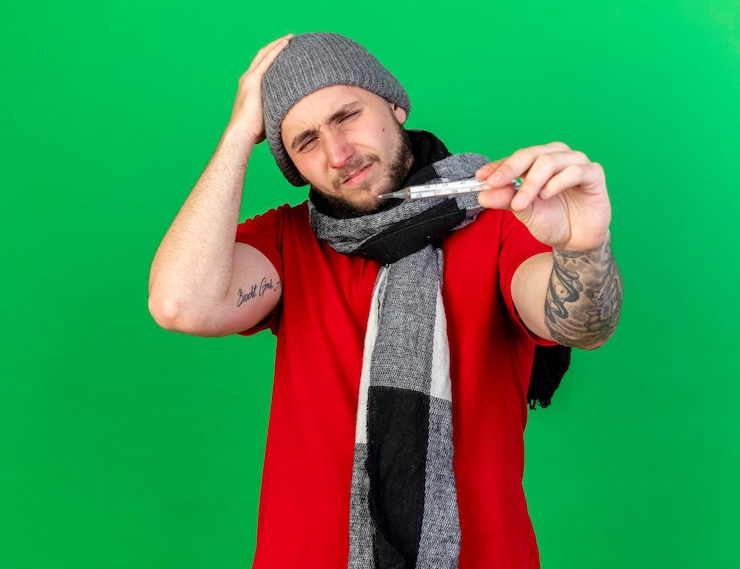 Foto gratuito dolor joven enfermo caucásico vistiendo gorro y bufanda de invierno pone la mano en la cabeza y sostiene el termómetro