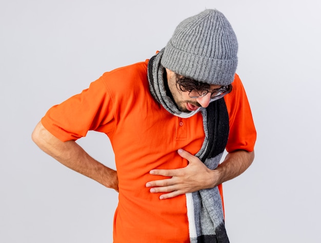 Dolor joven enfermo con gafas, gorro de invierno y bufanda poniendo las manos en el vientre y detrás de la espalda aislado en la pared blanca