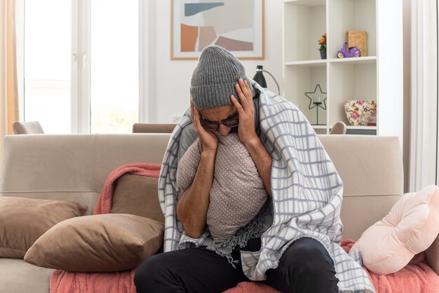 Dolor joven enfermo con gafas ópticas envuelto en cuadros con bufanda alrededor del cuello con sombrero de invierno poniendo las manos en la cabeza sosteniendo la almohada sentado en el sofá en la sala de estar