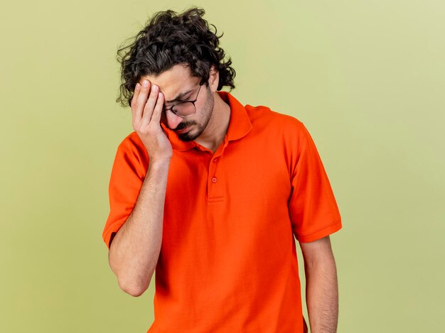 Dolor joven enfermo con gafas poniendo la mano en la cabeza con los ojos cerrados aislado en la pared verde oliva con espacio de copia