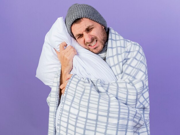 Dolor joven enfermo con sombrero de invierno con bufanda envuelta en cuadros y almohada abrazado aislado sobre fondo púrpura