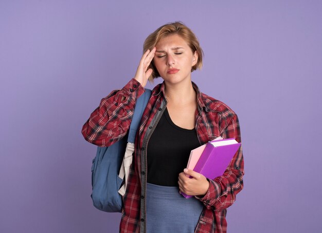 Dolor joven estudiante eslava vistiendo mochila poniendo la mano en la frente sosteniendo el libro y el cuaderno