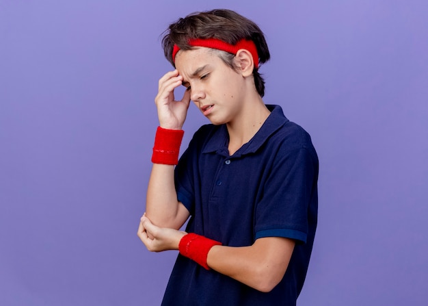 Dolor de joven guapo deportivo vistiendo diadema y muñequeras tocando la cabeza manteniendo la mano en el codo mirando hacia abajo sufriendo de dolor de cabeza aislado