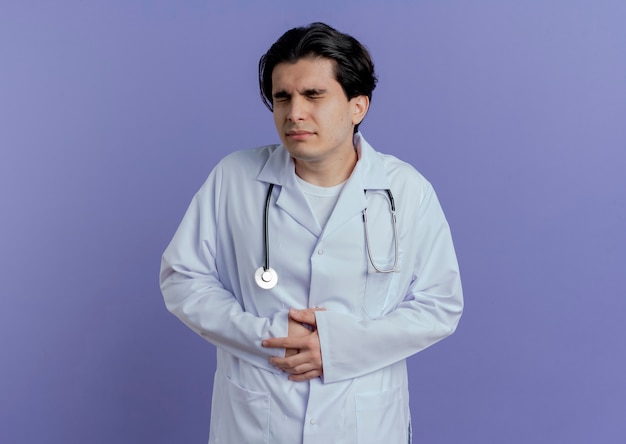 Dolor joven médico vistiendo bata médica y estetoscopio sosteniendo el vientre con los ojos cerrados aislado en la pared púrpura con espacio de copia