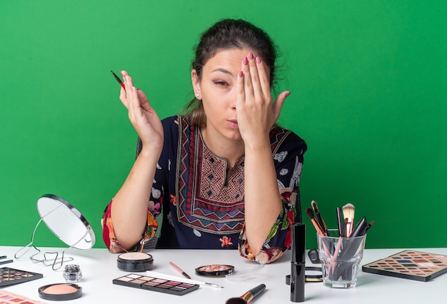 Dolor de joven morena sentada a la mesa con herramientas de maquillaje sosteniendo delineador de ojos y poniendo la mano en el ojo
