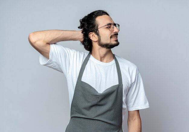 Dolor de joven peluquero masculino caucásico con gafas y banda de pelo ondulado en uniforme poniendo la mano detrás de la cabeza con los ojos cerrados