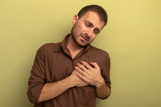 Dolor joven poniendo las manos en el corazón mirando hacia abajo aislado en la pared verde oliva