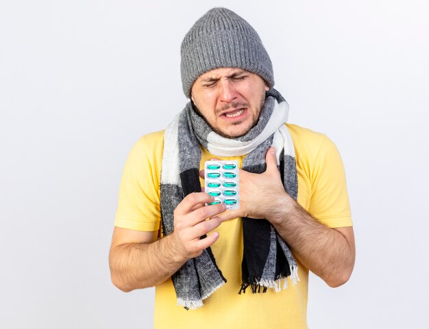 Foto gratuita dolor joven rubio enfermo con gorro de invierno y bufanda pone la mano en el pecho y sostiene un paquete de píldoras médicas aisladas en la pared blanca