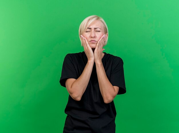 Foto gratuita dolor de mujer eslava rubia de mediana edad poniendo las manos en las mejillas que sufren de dolor de muelas con los ojos cerrados aislados en la pared verde con espacio de copia