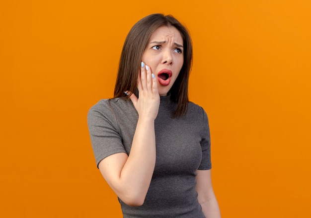 Dolor de mujer joven y bonita mirando directamente y poniendo la mano en la mejilla que sufre de dolor de muelas aislado sobre fondo naranja con espacio de copia