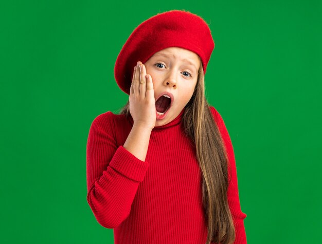Dolor de niña rubia vistiendo boina roja mirando al frente manteniendo la mano en la barbilla con la boca abierta aislada en la pared verde con espacio de copia