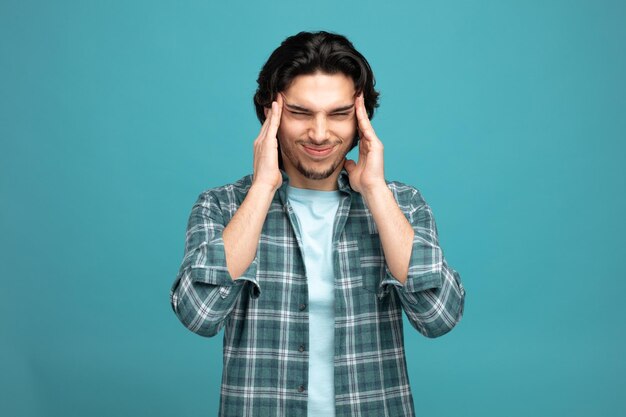 dolorido joven guapo manteniendo las manos en la cabeza con los ojos cerrados con dolor de cabeza aislado sobre fondo azul