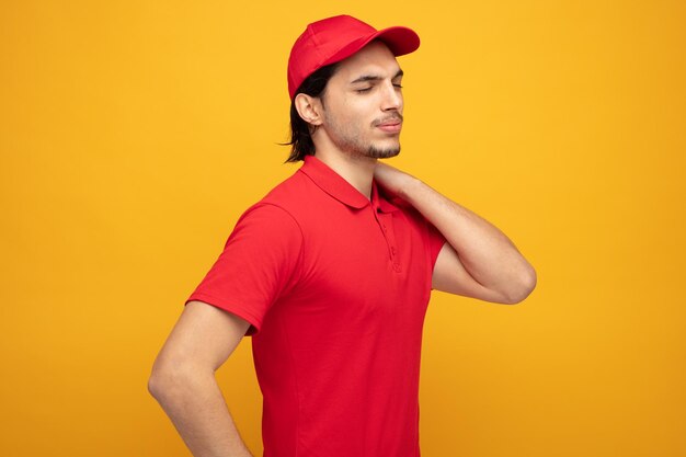 dolorido joven repartidor vestido con uniforme y gorra de pie en la vista de perfil manteniendo la mano en el cuello con los ojos cerrados aislado sobre fondo amarillo