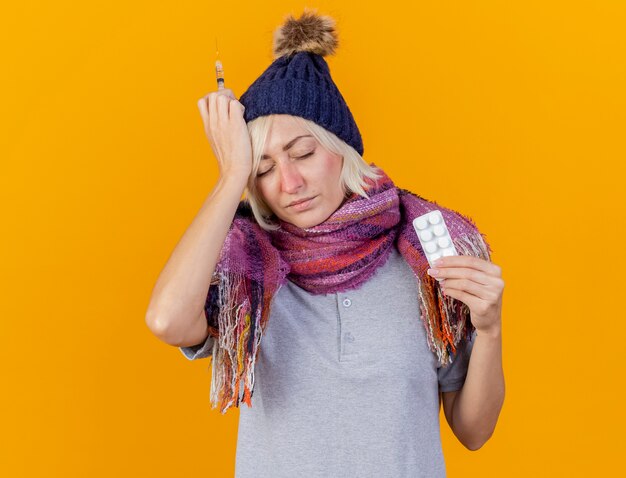 Dolorosa joven rubia enferma eslava con gorro de invierno y bufanda pone la mano en la cabeza sosteniendo la jeringa y el paquete de píldoras médicas aisladas en la pared naranja con espacio de copia