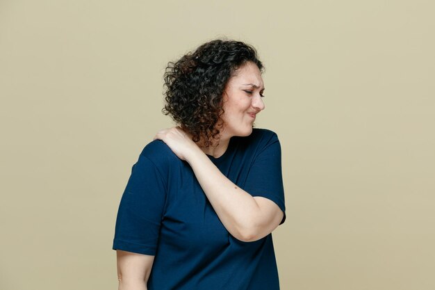 Dolorosa mujer de mediana edad con camiseta manteniendo la mano en el hombro con los ojos cerrados aislado en el fondo verde oliva