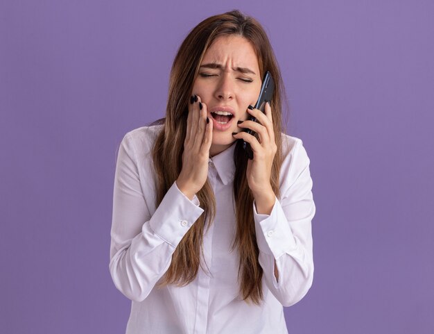 Doloroso joven bastante caucásica pone la mano en la cara y habla por teléfono aislado en la pared púrpura con espacio de copia