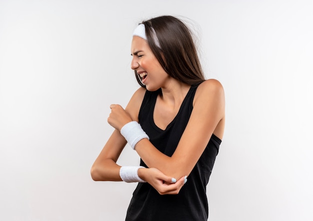 Foto gratuita doloroso joven bastante deportivo vistiendo diadema y muñequera poniendo la mano en el brazo con los ojos cerrados en la pared blanca con espacio de copia