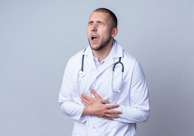 Doloroso joven médico con bata médica y un estetoscopio alrededor de su cuello poniendo la mano en el vientre y otra en la muñeca con los ojos cerrados aislados en blanco con espacio de copia