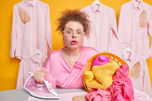 La doncella de pelo rizado emocional aturdida lleva una cesta de ropa sucia con detergentes ocupada haciendo planchado lleva glsses transparentes y poses de bata contra camisas colgadas en perchas. Tareas domésticas