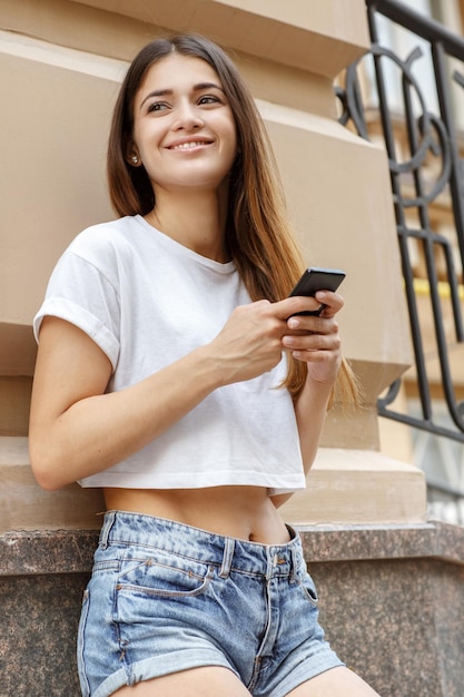 Dónde ir a continuación Retrato de medio cuerpo de una joven y hermosa morena parada cerca de la pared mirando alrededor y sosteniendo su celular