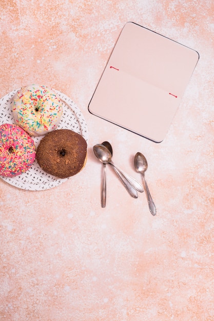 Donuts surtidos con chocolate glaseado; rosa glaseada y asperja donas en un plato blanco y una libreta en blanco sobre un fondo rústico