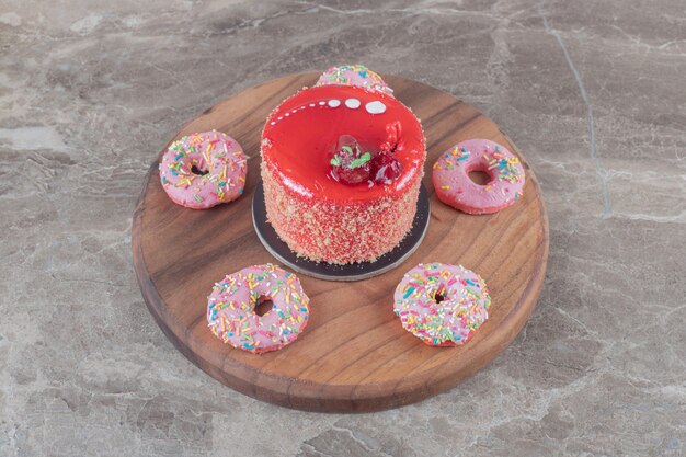 Donuts del tamaño de un bocadillo alrededor de un pastel cubierto con jarabe de fresa en una tabla sobre una superficie de mármol