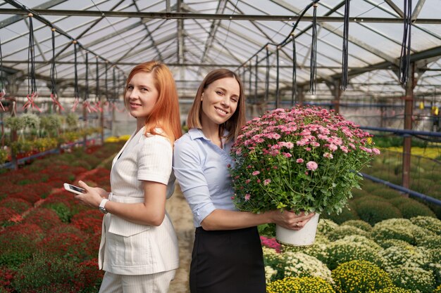 Dos adorables damas posando con un ramo de crisantemos rosados en una hermosa casa verde en flor con techo de cristal.